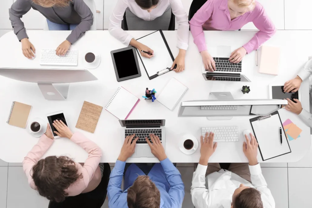 Overhead view of a group of people working around a table with computers, tablets, and notes.