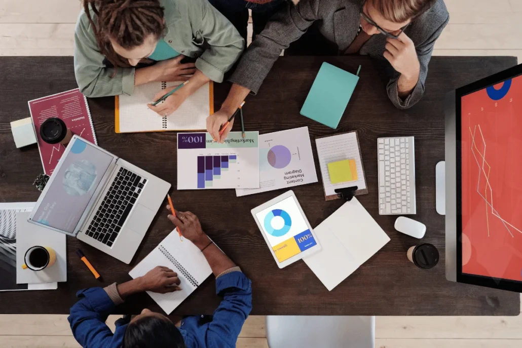 Top-down view of four people collaborating at a table with laptops, charts, and notes.