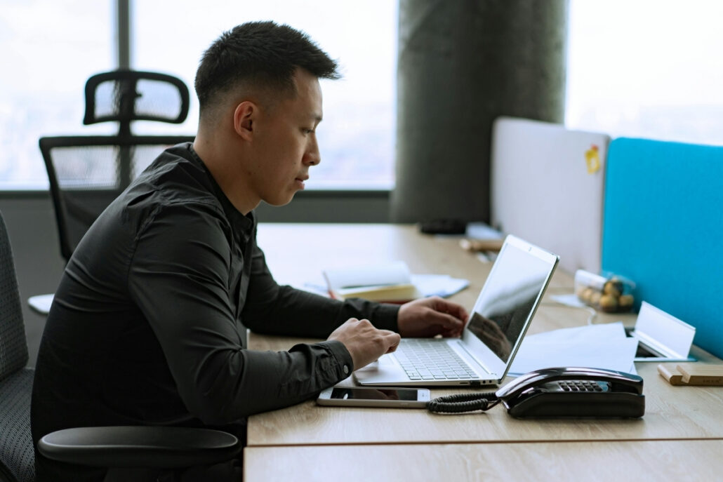 Man in a black shirt working on a laptop in a modern office with a phone on the desk.