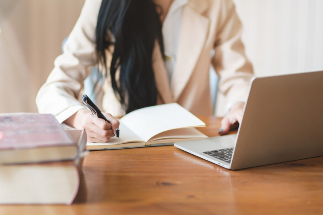 Person writing in notebook next to laptop and books on a desk.