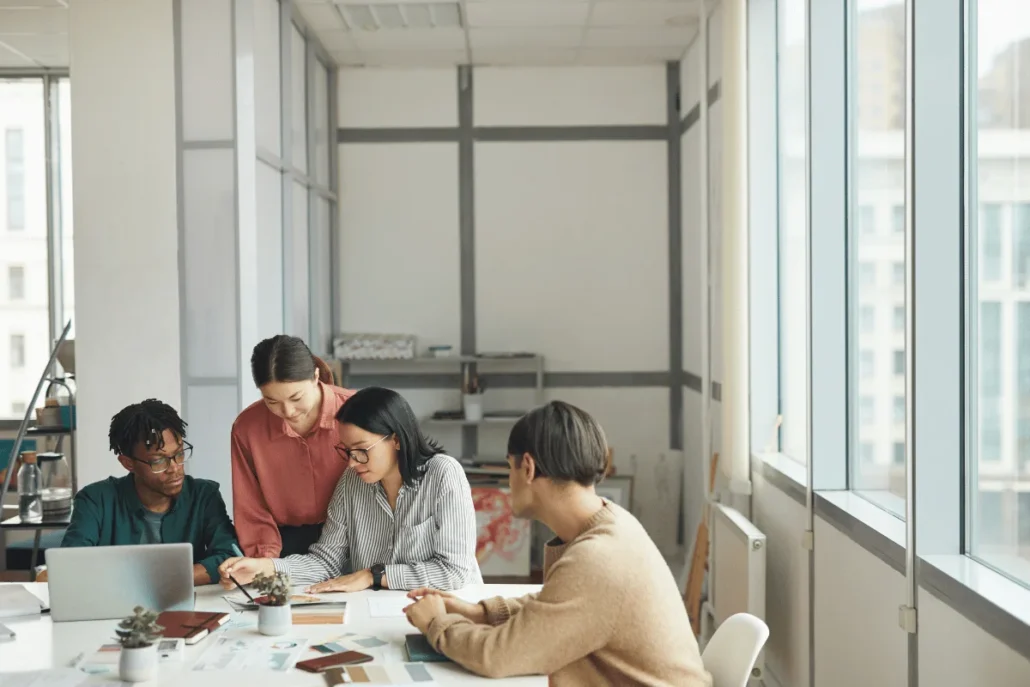 Four people in a meeting with laptops and papers on a table in a bright office setting.