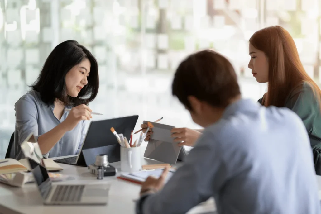 Three people at a table with digital tablets and a laptop in a bright office.