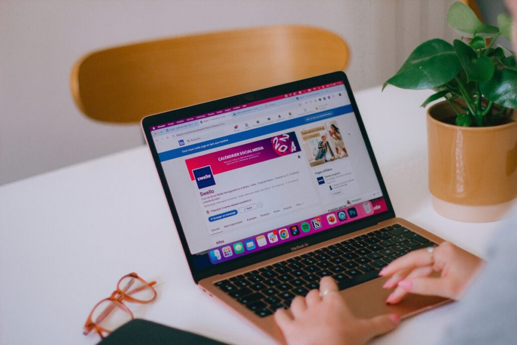 Person using a laptop with a social media site on the screen, glasses on the table, and a plant beside.