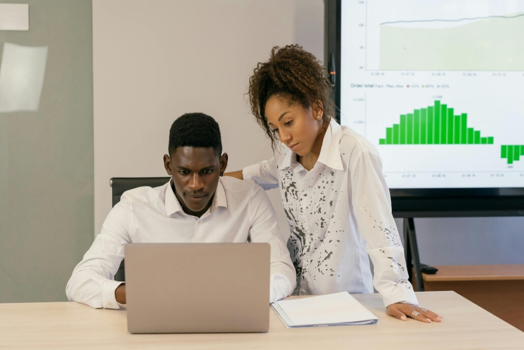 Two professionals at a table with a laptop and graphs on a screen in the background.