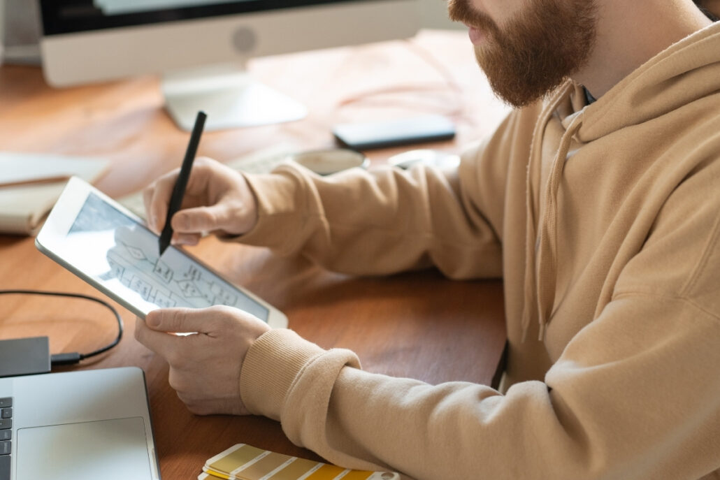 Person with a beard drawing on a tablet with a stylus, with computer and smartphone nearby.