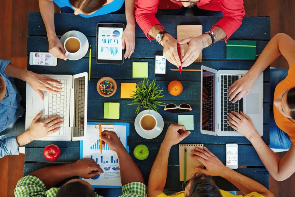 Overhead view of a busy team meeting with laptops, tablets, and paperwork on a table.