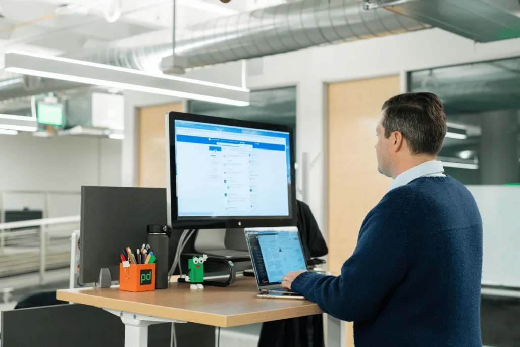 Man in office standing by desk with a monitor and laptop, looking at the screen.