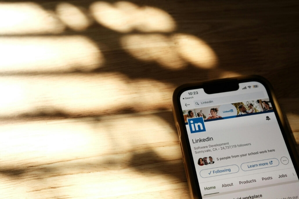 Smartphone on a wooden surface displaying the LinkedIn app, dappled sunlight casting shadows.