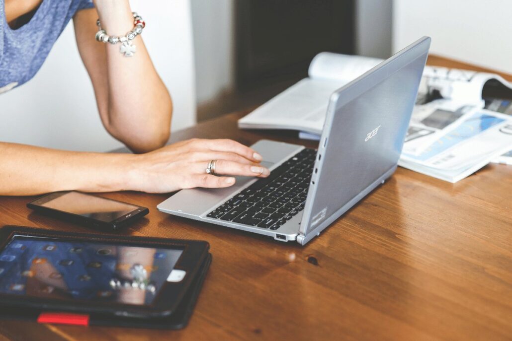 Person typing on a laptop with a tablet and smartphone on a wooden desk.