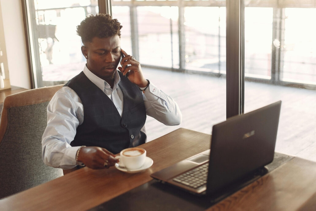 A man in a vest and tie sits at a table, working on a laptop with a cup of coffee beside him.