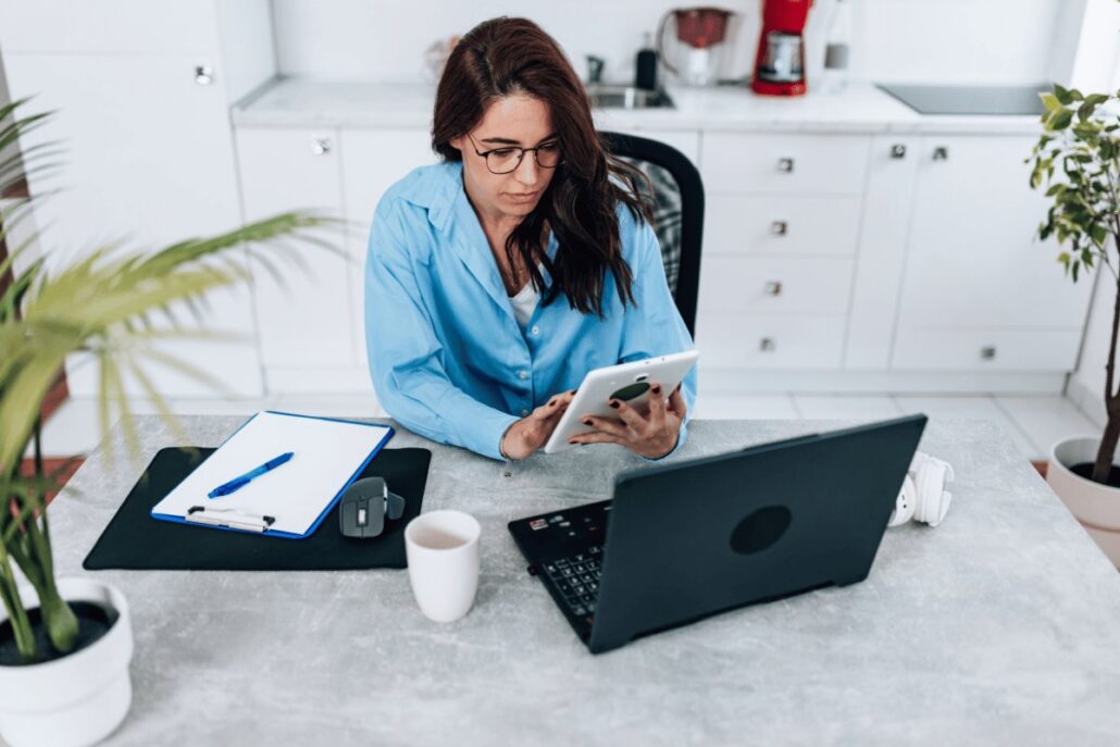 Person in blue shirt using a tablet at a desk with a laptop, clipboard, and coffee cup.