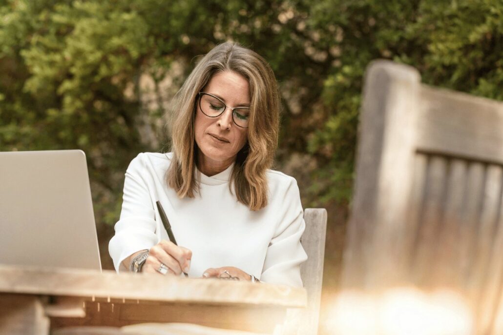 Woman in white outfit writing in a notebook at an outdoor table with a laptop beside her.