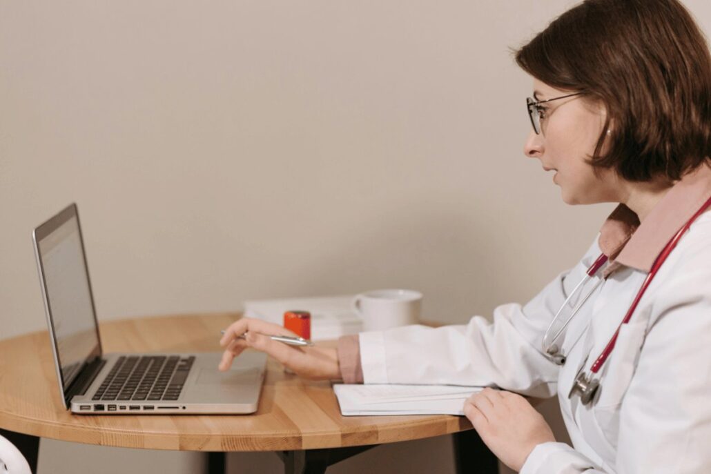 Medical professional in a lab coat using a laptop at a wooden desk, with a notebook and a coffee cup.