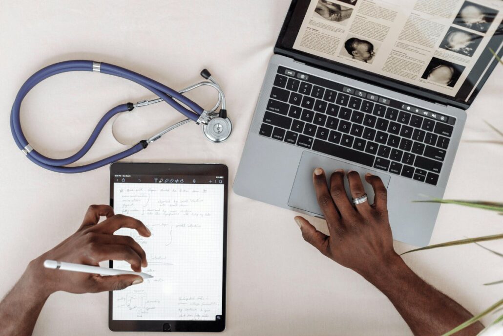 Person studying medical notes on tablet beside laptop with stethoscope on desk.