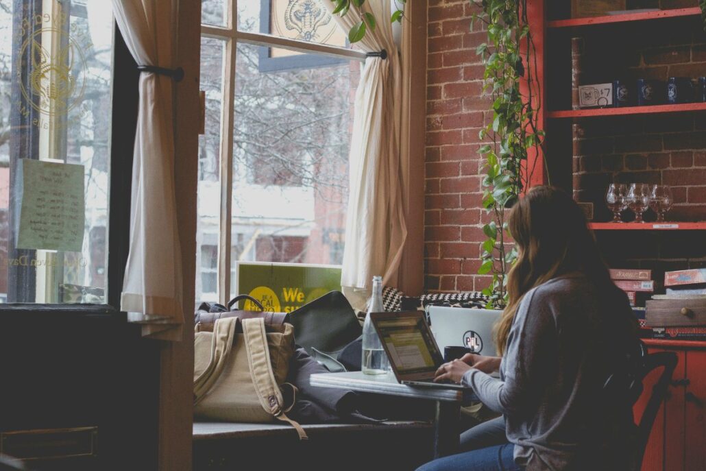 Woman working on a laptop in a cozy cafe with brick walls and a large window.