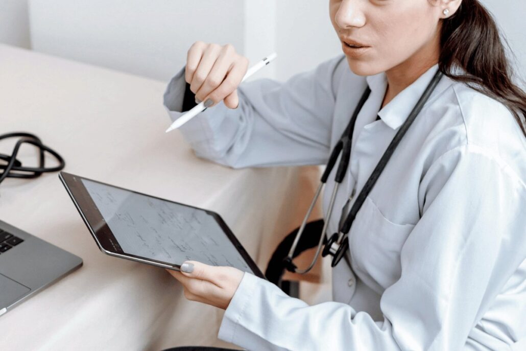 Professional in a white coat using a digital tablet with a stylus next to a laptop on a desk.