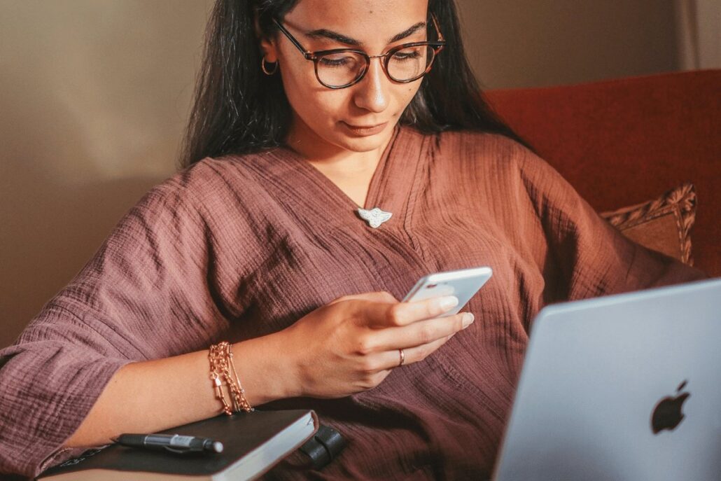 A woman wearing glasses is sitting on a couch, focused on her phone.