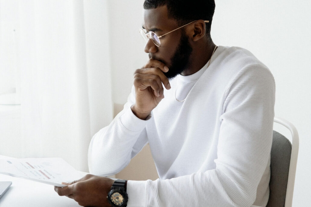 Man in white sweater holding a document, seated at a white table with a laptop.