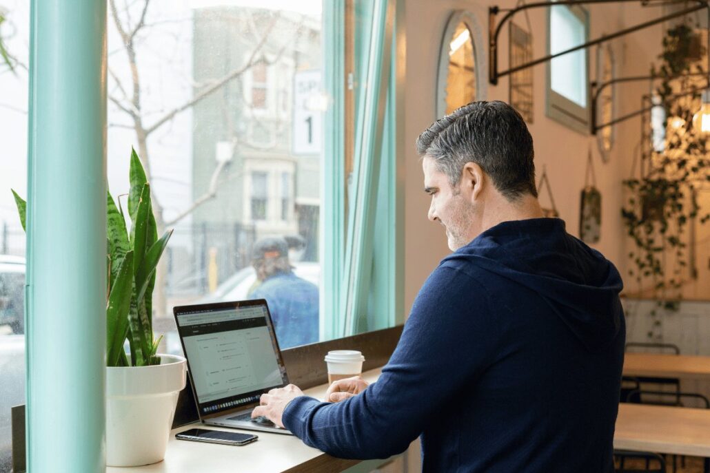 Man working on a laptop in a cozy cafe near a window, surrounded by greenery and natural light.