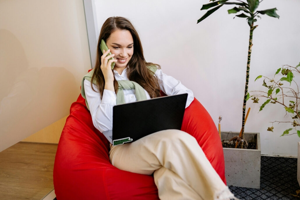 An individual sitting on a red bean bag using a laptop and holding a phone.