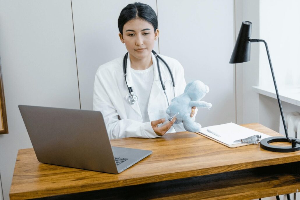 Doctor in white coat with stethoscope holding a toy, laptop, and notes on desk.