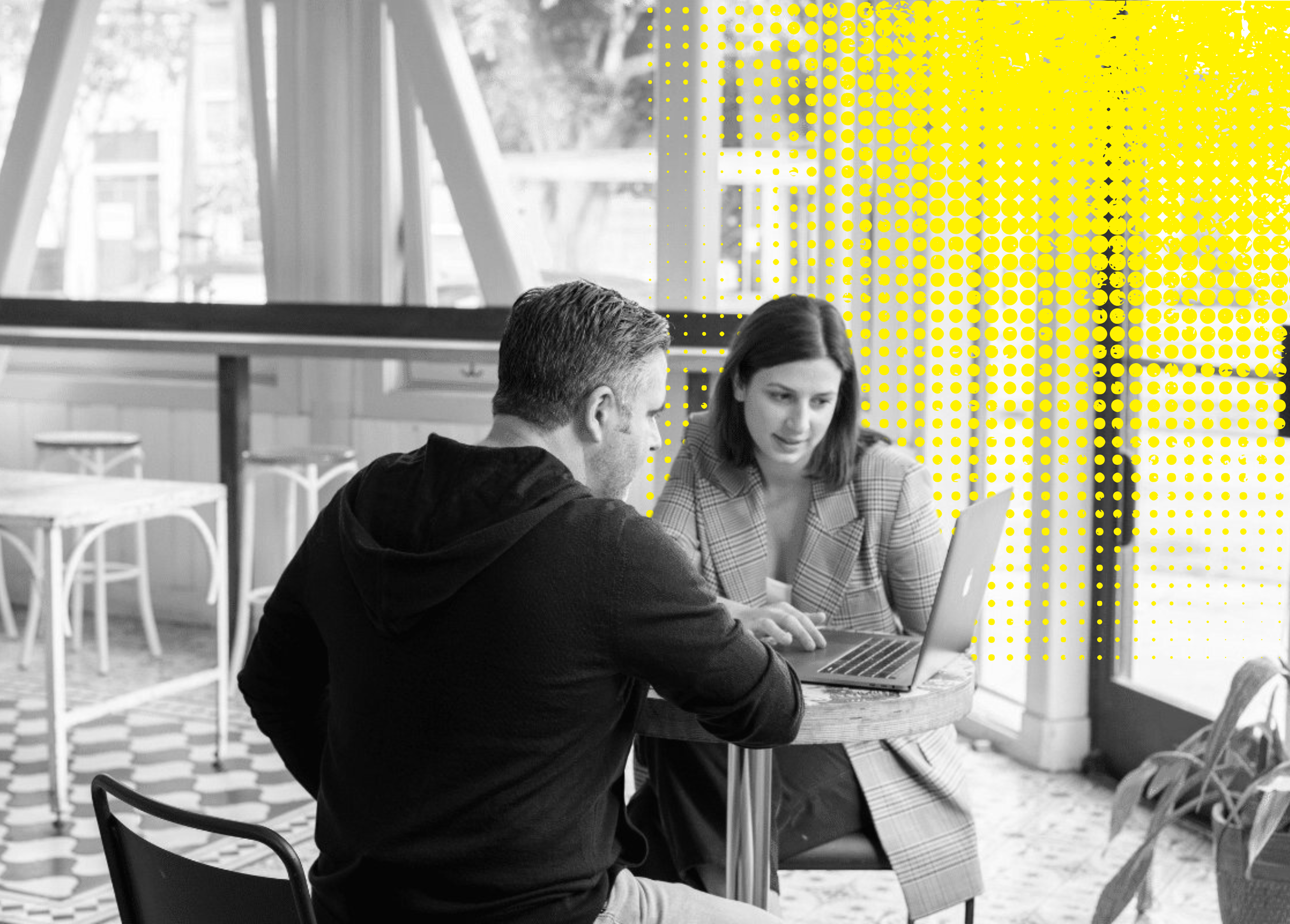 Two individuals sitting at a table with one using a laptop in a modern cafe setting.