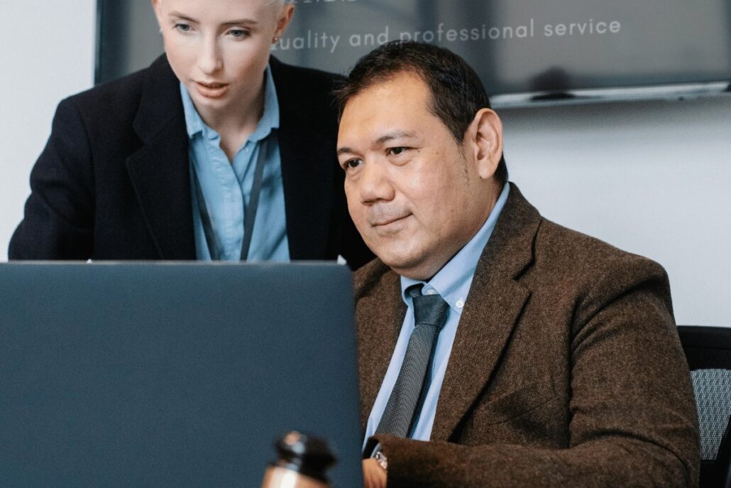 Two professionals in a meeting room with a laptop and presentation screen.