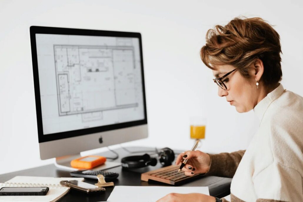 A person working on an architectural plan on a computer with stationery and a glass of juice nearby.