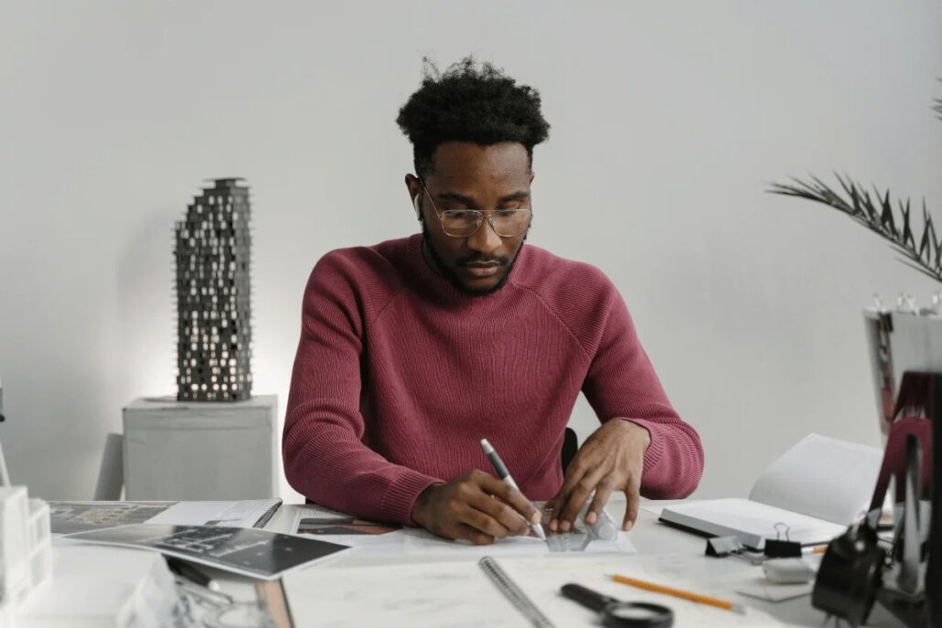 Person in a pink sweater writing at a desk with papers and a model structure.