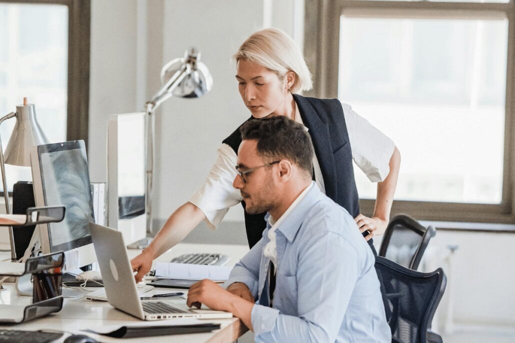 Two professionals collaborating at an office workstation with computers and phones.