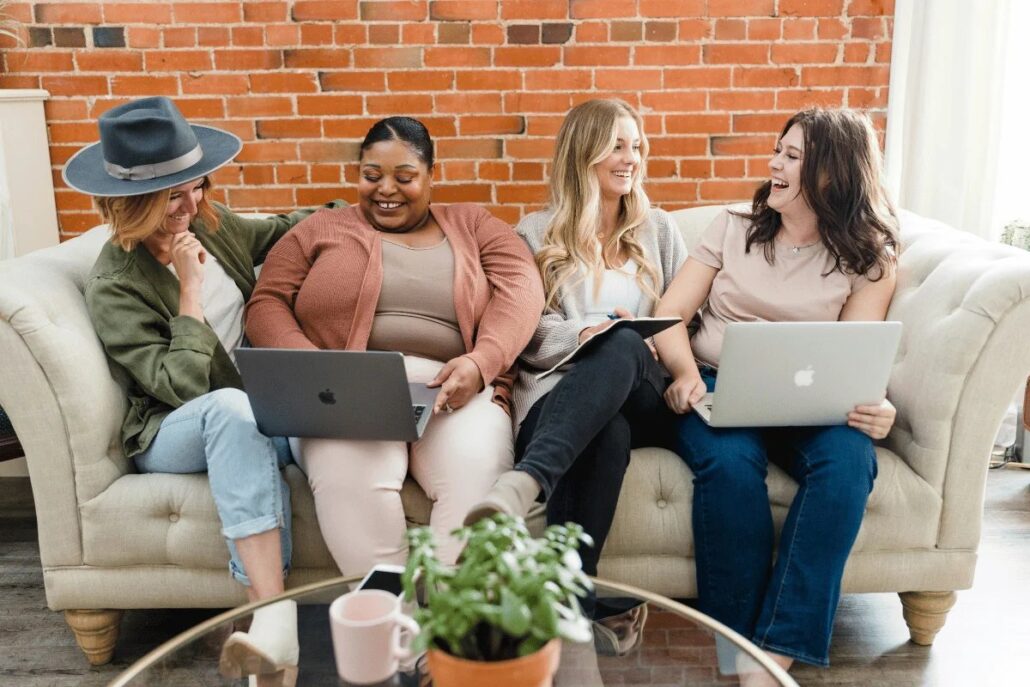 Four people sitting on a sofa with laptops, brick wall background.