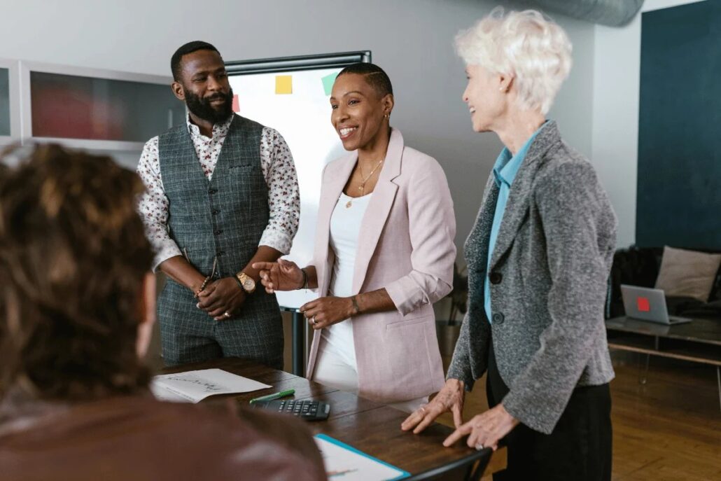 Three professionals shaking hands in a meeting room, standing by a table with papers.