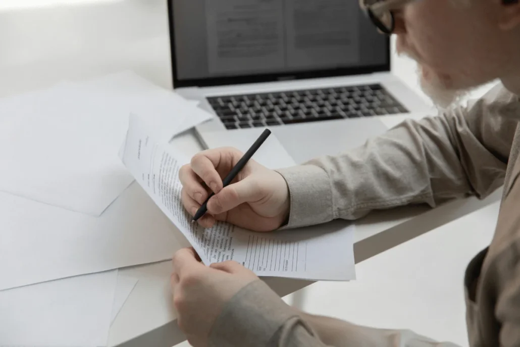 Person reviewing documents at a desk with a laptop in the background.