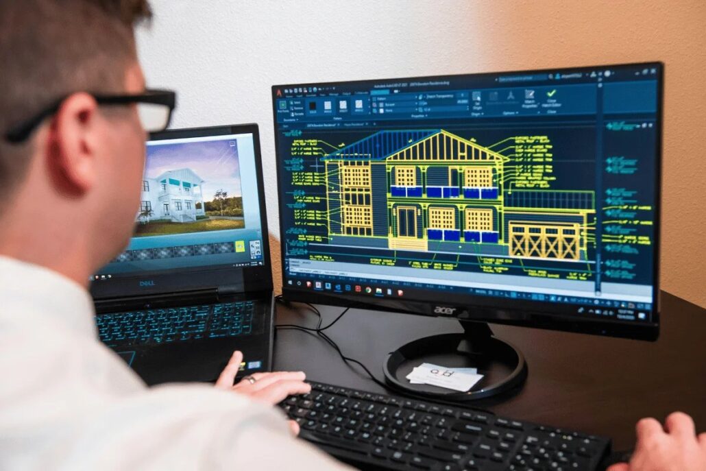 Person at desk viewing CAD house design on dual monitors.