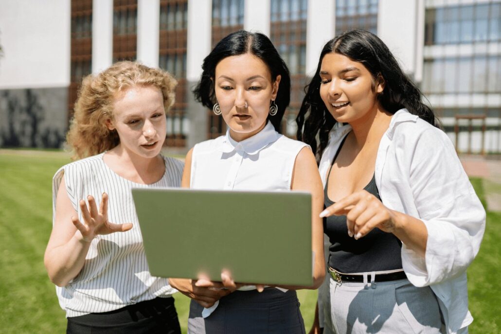 Three individuals stand outside holding a laptop, engaged in a discussion.