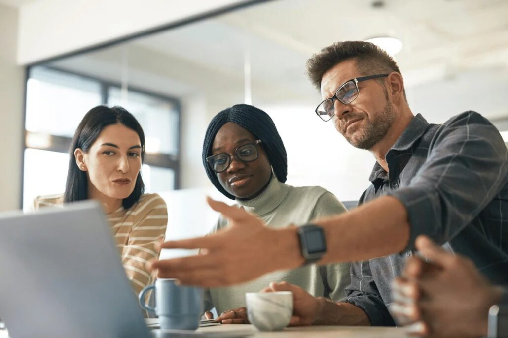 Three colleagues in a discussion at a table with a laptop and a coffee cup.