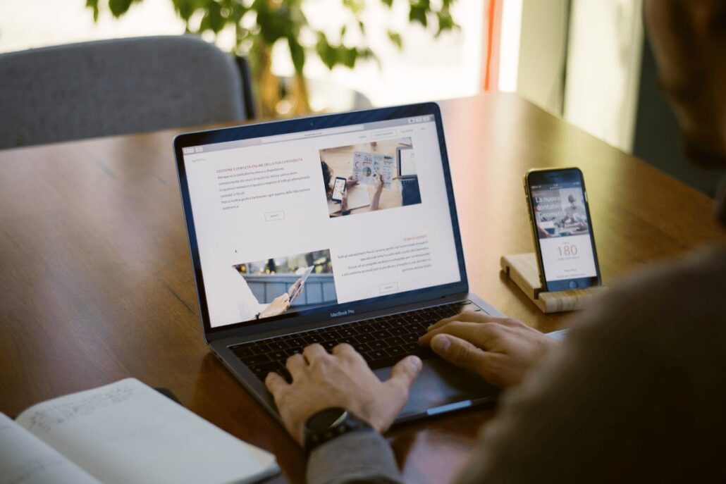 Person working on a laptop with a smartphone beside it, on a wooden desk with a notepad.