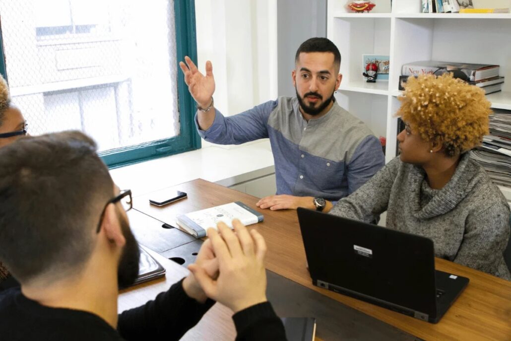 Three people in a meeting with laptops and a smartphone on a wooden table.