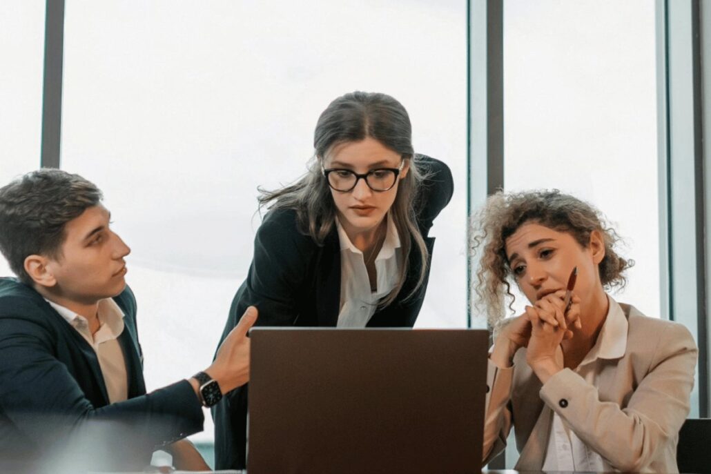 Three professionals in a meeting with a laptop by a window.