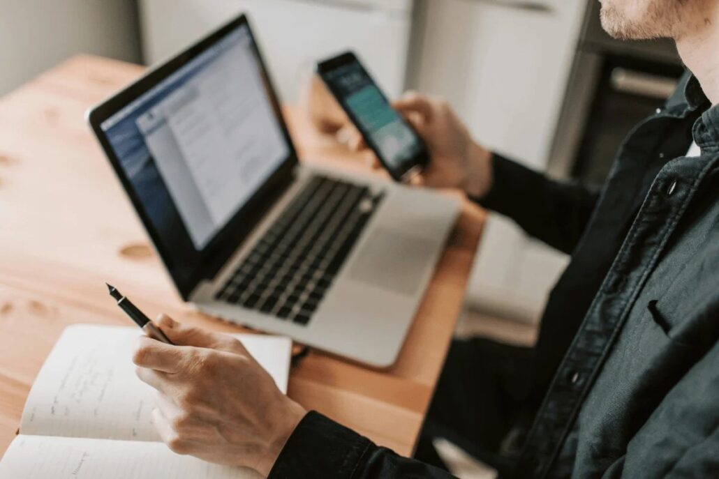 A professional sits at a wooden desk, using a laptop and smartphone, jotting notes in a notebook with a pen.