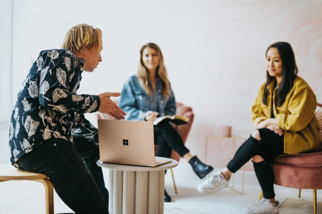 A group discussion in a stylish room, featuring a laptop on a round table and three individuals engaged in conversation.