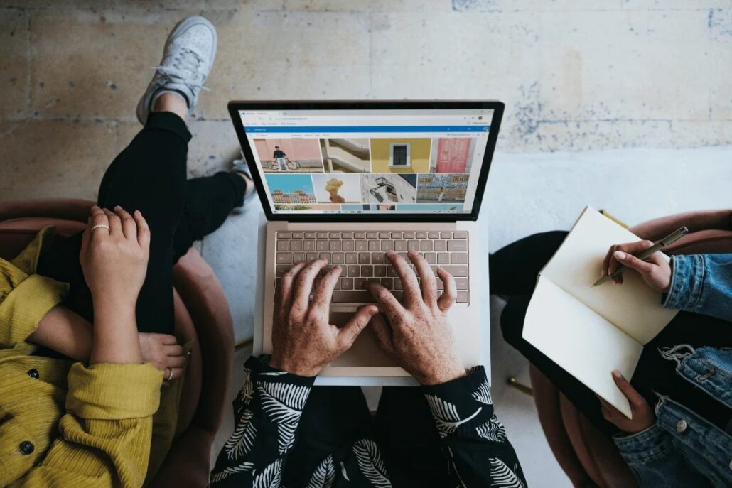 Aerial view of hands typing on a laptop displaying colorful images, with two people nearby holding a notebook and pen.