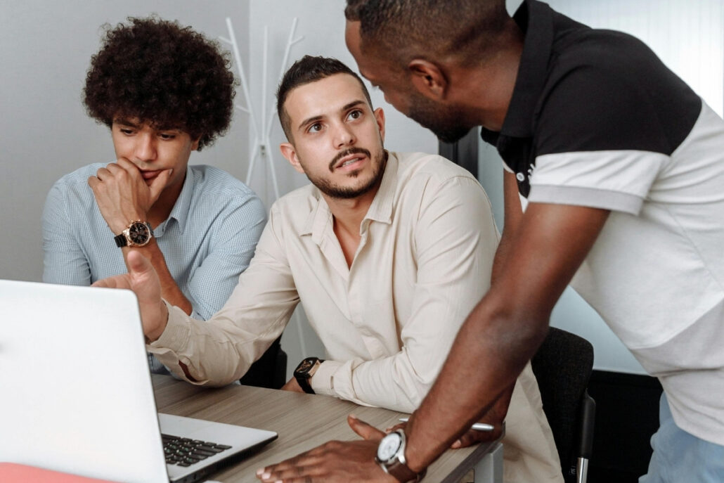 Three colleagues engaged in a focused discussion around a laptop in an office setting.