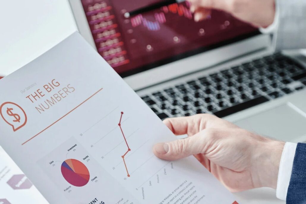 A hand holds a report titled "The Big Numbers," featuring graphs and charts, alongside a laptop displaying financial data analysis.