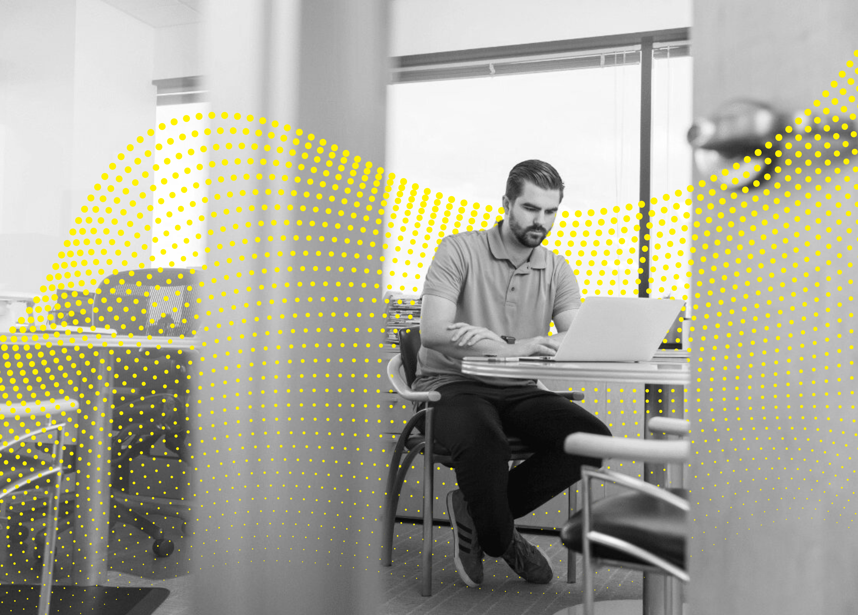 A person sits at a table in an office, working intently on a laptop with a bright, dotted design overlay in the corner.