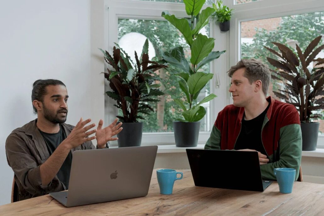 Two people engage in discussion at a wooden table adorned with laptops, surrounded by greenery in a bright, modern office.