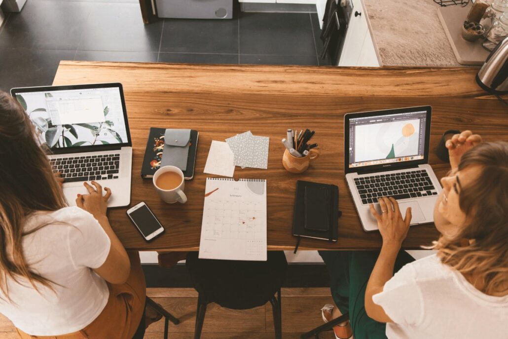 Two people working on laptops at a wooden table, surrounded by a calendar, notebooks, and coffee cups in a cozy workspace.
