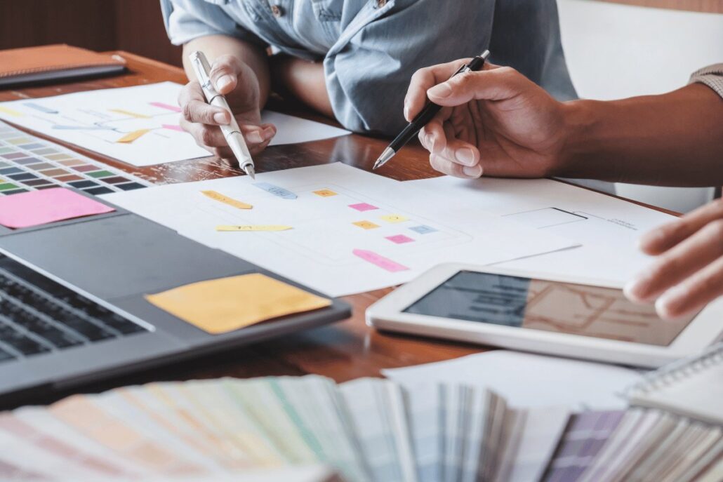 Two designers working on UI/UX wireframes and user flow diagrams, with color swatches and post-it notes spread across a desk.