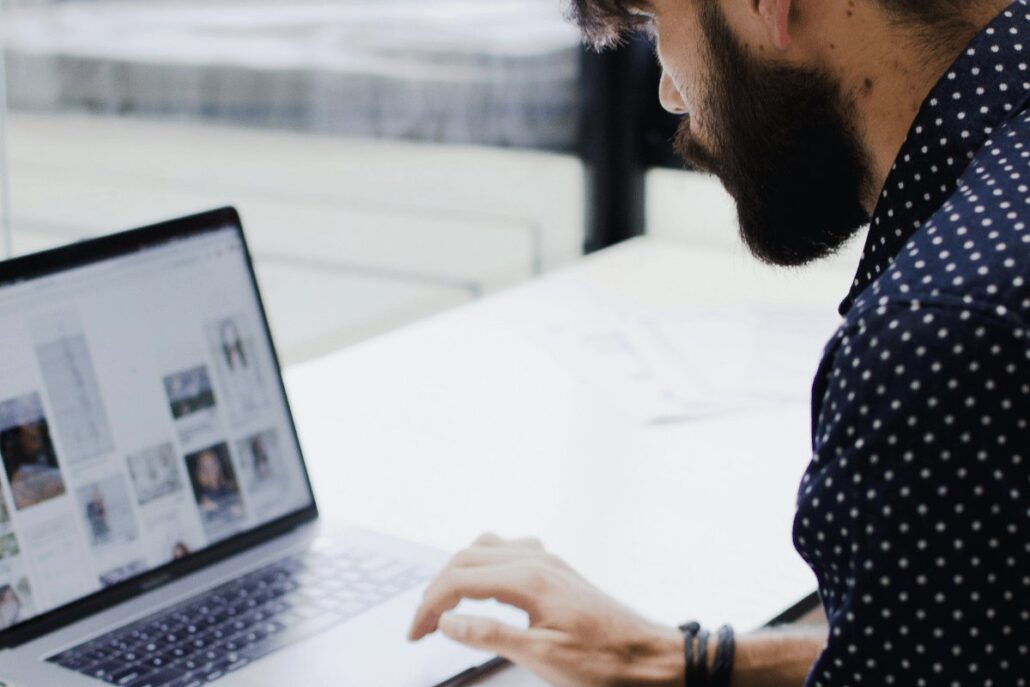 A person with dark hair is typing on a laptop, browsing images, while sitting at a white desk in a bright, minimalistic environment.