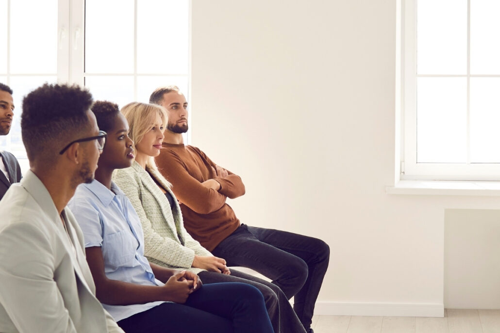 A diverse group of people sitting in a bright room, gazing towards a window, suggesting anticipation or engagement in discussion.